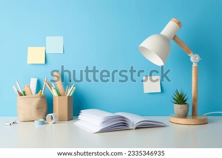 Side view photo of desk supplies for school: pencil organizers, pens rulers, open notebook, lamp, flowerpot with plant, sticky notes, and tape. Blue wall backdrop, perfect for text or ad integration