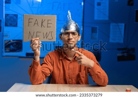 Male conspiracy theorist in a protective foil cap and glasses debunks myths holding poster fake news. Conspiracy theory concept. The schizophrenic works. Copy space. Royalty-Free Stock Photo #2335373279