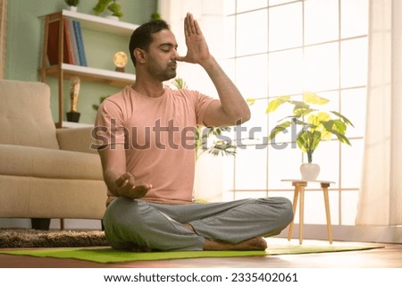 Peaceful indian man doing nostril breathing or pranayama yoga by closing eyes during morning at home - concept of healthy lifestyle, wellness and mindfulness Royalty-Free Stock Photo #2335402061