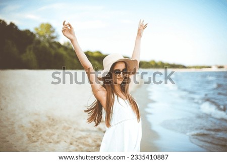 Happy smiling woman in free happiness bliss on ocean beach standing with a hat, sunglasses, and rasing hands. Portrait of a multicultural female model in white summer dress enjoying nature during Royalty-Free Stock Photo #2335410887