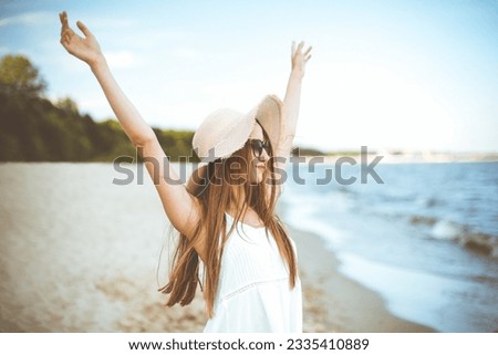 Happy smiling woman in free happiness bliss on ocean beach standing with a hat, sunglasses, and rasing hands. Portrait of a multicultural female model in white summer dress enjoying nature during Royalty-Free Stock Photo #2335410889