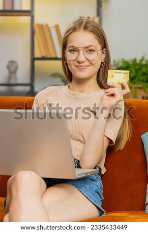 Young blonde woman using credit bank card and laptop computer notebook while transferring money, purchases online shopping, order food delivery at home. Adult girl in room sitting on couch. Vertical
