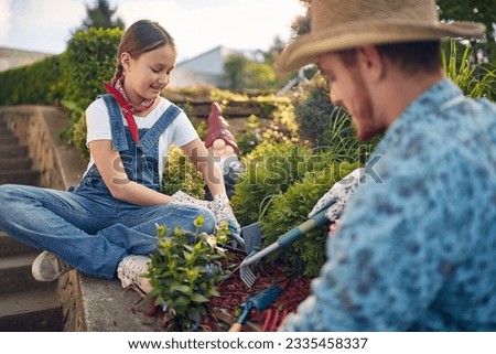 Beautiful young girl using garden rake sitting by the garden outdoors planting flowers with her father on a sunny summer day. Home, lifestyle, family concept. Royalty-Free Stock Photo #2335458337