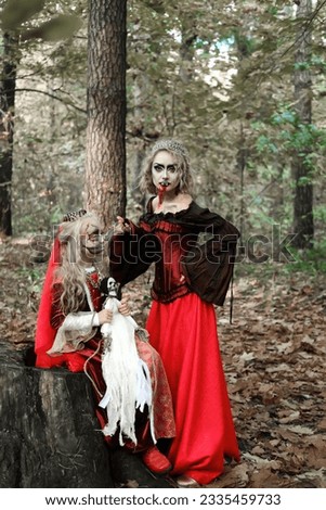 A woman in the form of a vampire or a sorceress and a girl in the form of a dead princess with a skeleton doll in their hands pose in the forest. Models are looking at camera. Vertical photo