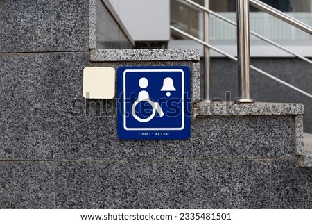 The blue logo of people with disabilities next to the button to call for help and braille text in front of the gray steps at the entrance to the building