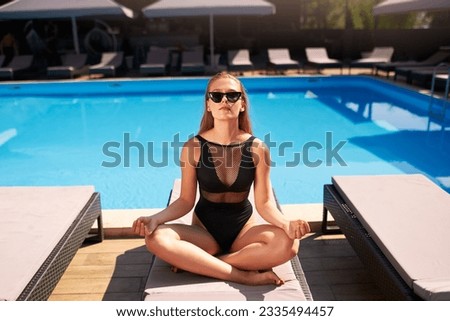 Young athletic woman in bikini meditating by the swimming pool at tropical spa. Fit girl in one piece swimwear practicing zen yoga in resort. Peaceful slim female sitting in lotus pose on beach bed.