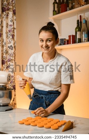 Young smiling woman with confectionery bag squeezing cream filling to macarons shells at kitchen interior. Homemade bakery concept