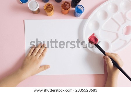 Top view of kid hands painting a colorful picture with a paintbrush, acrylic paint colors in tubes, and color palette on pink pastel background. School supplies, preschool child painting.