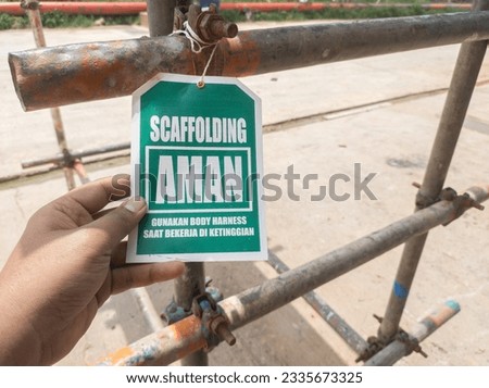 Hanged green sign label tag with writing in scaffolding aman, gunakan body harness saat bekerja di ketinggian in English safe scaffolding, use body harness when working at heigh

