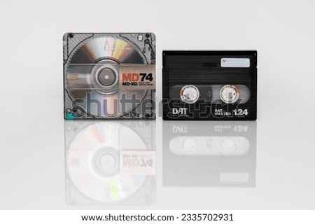 A minidisc shimmering with iridescent reflections and a DAT cassette stand side by side vertically isolated on a white background with reflection