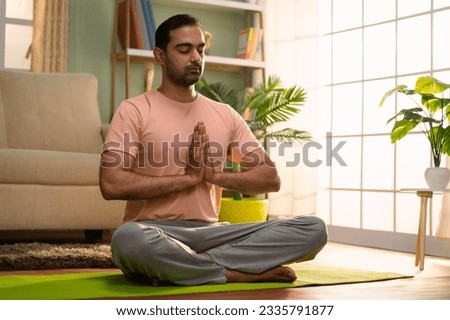 Indian young man doing namaste posture or yoga with closed eyes while sitting at home - concept of healthy lifestyle, fitness and self caring Royalty-Free Stock Photo #2335791877