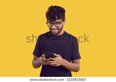 Young joyful Indian man chatting with phone and reading SMS with smile using instant messengers and social networks standing in yellow studio. Mobile technologies, internet communications Royalty-Free Stock Photo #2335815607