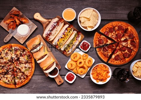 Junk food table scene. Pizza, hamburgers, chicken wings, hot dogs and salty snacks. Above view over a dark wood background. Royalty-Free Stock Photo #2335879893