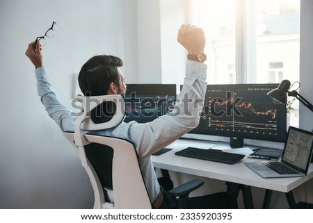 Yes Back view of stock trader with raised hands looking at multiple computer screens with data and charts and feeling happy while sitting in modern office Royalty-Free Stock Photo #2335920395