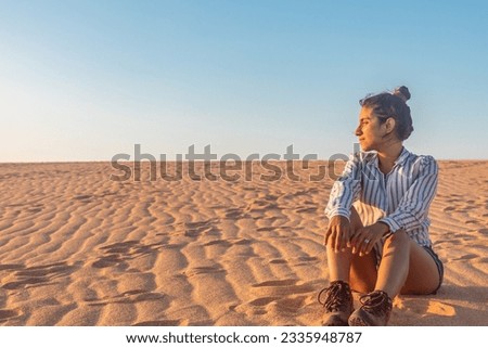 Captivating high desert sunset: Latina woman sitting, embracing the warm glow, surrounded by majestic dunes in the Guajira desert Royalty-Free Stock Photo #2335948787