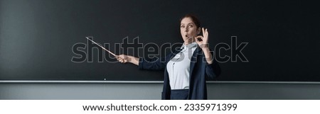 Red-haired caucasian woman in a pantsuit shows an ok sign. Surprised female teacher with a pointer at the blackboard. Widescreen. 