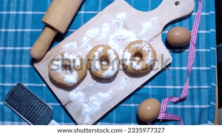 Donuts sprinkled with white powdered sugar on a wooden cutting board. You can also see tools and ingredients for making donut cakes. Donuts are a craze for young and old alike