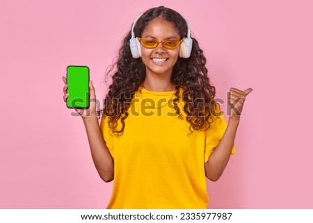 Young happy Indian woman teenager in wireless headphones holding mobile phone and smiling looking at camera demonstrating green screen of gadget stands on pink background. Music playlist concept