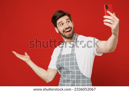 Young fun male housewife housekeeper chef cook baker man wear grey apron do selfie shot on mobile cell phone post photo point hand aside on area isolated on plain red background. Cooking food concept