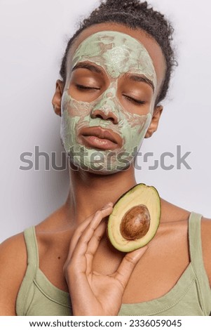 Calm young dark haired African woman indulges in self care applying beauty mask with avocado in hand wants to have radiant skin keeps eyes closed wears tshirt isolated over white background.