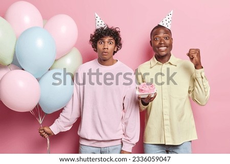 Happy smiling broadly African american and surpirsed Hindu guys celebrate birthday having party holding cake with candles and bunch of colourful balloons standing on pink background in centre