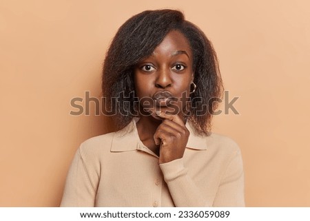 People emotions concept. Studio photo of young pretty thoughtful African american woman standing in centre isolated on beige background keeping right hand raised under chin looking straight at camera