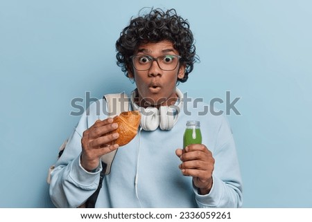 People emotions concept and student lifestyle. Indoor photo of young surprised Hindu male as if seeing something unusual or unexpected holding delicious croissant and small bottle of green smoothie