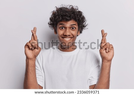 Cheerful Hindu man with curly hair stands with hopeful expression demonstrating his eager anticipation keeps fingers crossed dressed in casual t shirt isolated on white wall. May dreams come true Royalty-Free Stock Photo #2336059371