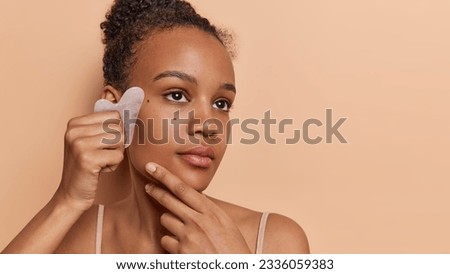 Serious teenage girl using gua sha stone skillfully massages and lifts her face enhancing natural beauty undergoes beauty procedures at home poses indoor concentrated aside isolated over brown wall