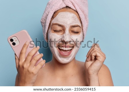 Photo of joyful woman applies nourishing beauty mask her glowing smile reflecting inner joy and contentment exclaims loudly and clenches fist holds mobile phone stands shirtless isolated on blue wall