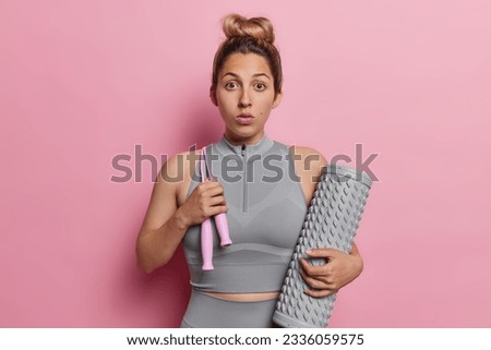 Horizontal shot of impressed young European woman carries massage roller and jumping rope feels stunned goes in for sport regularly reacts to something shocking isolated over pink background