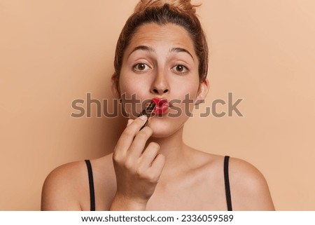 Beauty and cosmetic. Studio shot of young pretty glad European lady applying red lipstick standing in centre isolated on beige background looking straight at camera keeping long brunette hair in bun