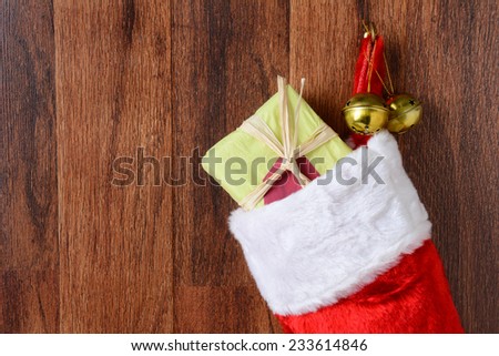 Closeup of a Christmas stocking filled with presents hanging from a hook on a wood wall. Two jingle bells also hang from the hook in horizontal format.