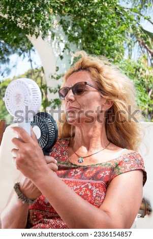 Heatwave image of a woman with two handheld fans blowing on her face as she struggles with the searing heat. Selective focus on the woman's glasses.