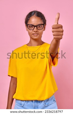 Young attractive ethnic Indian millennial woman showing thumbs up to praise advertised product and service dressed in yellow t-shirt and jeans stands on isolated pink background. Teenager, schoolgirl