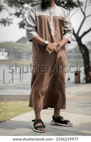 young asian man posing with wearing brown modern style thawb standing on outdoor during sunny day. modern moslem outfit. fashion and lifestyle Royalty-Free Stock Photo #2336219977
