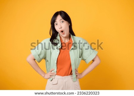 Expressive and surprising, a young 30s Asian woman wearing a green shirt on an orange shirt showing surprise face while standing with akimbo arms. unexpected with vibrant image on yellow background.