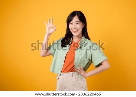 young Asian woman 30s, dressed in orange shirt and green jumper. Her okay hand gesture and gentle smile, isolated on a yellow background, convey a positive message through body language. Royalty-Free Stock Photo #2336220465