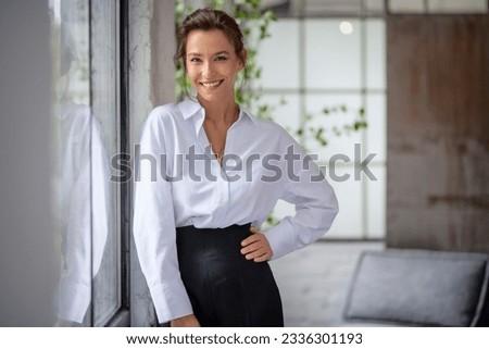 Portrait of a brown haired middle-aged woman wearing a white shirt and black pants and standing by the window. Attractive female looking at camera and smiling.  Royalty-Free Stock Photo #2336301193