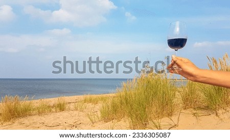 A womanly hand holding a glass filled with red wine  on a sandy beach by Baltic Sea on Sobieszewo island, Poland. The beach is scarcely overgrown with high grass. The sea is in the back. Celebration Royalty-Free Stock Photo #2336305087