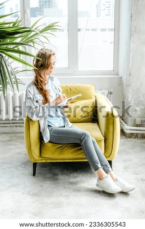 Young beautiful long haired college student girl in casual shirt with notebook,organizer pen sitting on yellow chair. Businesswoman making notes. Freelancer,remote job. Records in psychological diary.