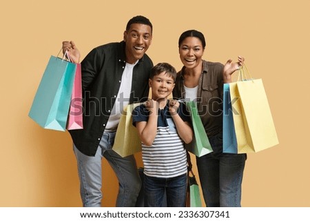 Cheerful cute young african american family of three father mother and preteen child son holding colorful purchases paper bags, have shopping together, isolated on beige background. Retail, shopping Royalty-Free Stock Photo #2336307341