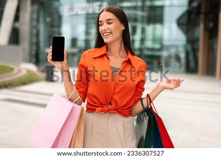 Shopping App. Cheerful Customer Woman Showing Cellphone With Blank Screen Standing With Shopper Bags In Urban Area. Shopaholic Lady Recommending Mobile App And Ecommerce Offer. Mockup