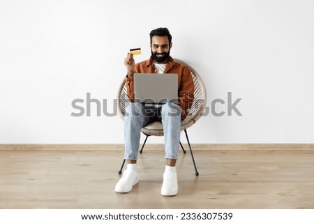 Excited indian man holding credit card and laptop computer, sitting on chair, shopping online against white studio wall indoor. Buyer buying things on internet web store posing with pc and bankcard