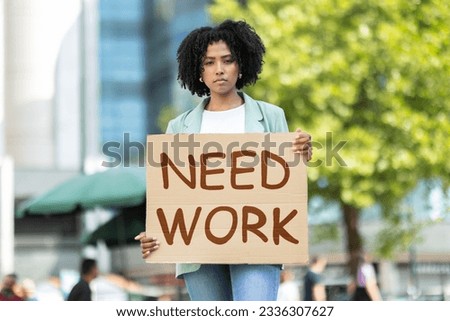 Unemployed pretty young black woman wearing casual outfit standing on street, holding Need Work placard, upset unhappy african american lady attending unemployment strike Royalty-Free Stock Photo #2336307627