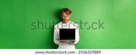 Impressed redhead nerdy guy in glasses showing blank laptop screen and looking at display amazed, standing over green background.