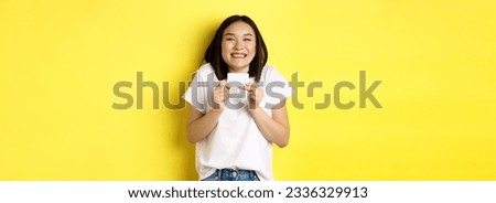 Young asian woman in casual white t-shirt going on shopping, showing plastic credit card and smiling excited, standing over yellow background.
