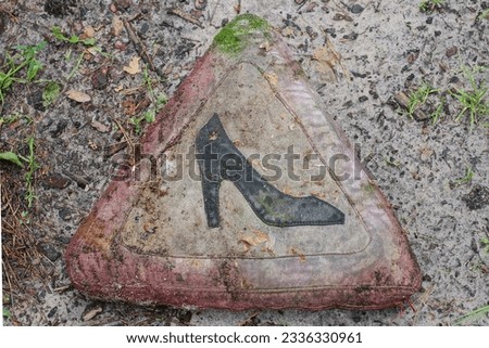 one old dirty triangular traffic sign a woman driving with an embroidered black shoe on a fabric on a pillow lies on the ground and green grass on the road in the street