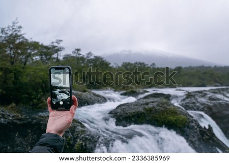 A hand holding a phone while taking a picture of the view in the park Saltos de Petrohue, in the south of Chile.