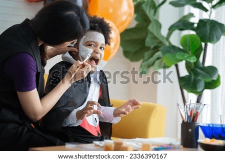 Mother and son in Halloween costumes having fun with makeup, mother paints a pattern on her son's face with a paintbrush, boy makes a frightening look at the camera